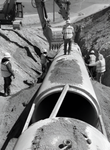 Workers guiding a large pipe section into a trench with heavy machinery.