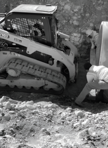 Construction worker inspecting a compact loader pressed against a trench wall.